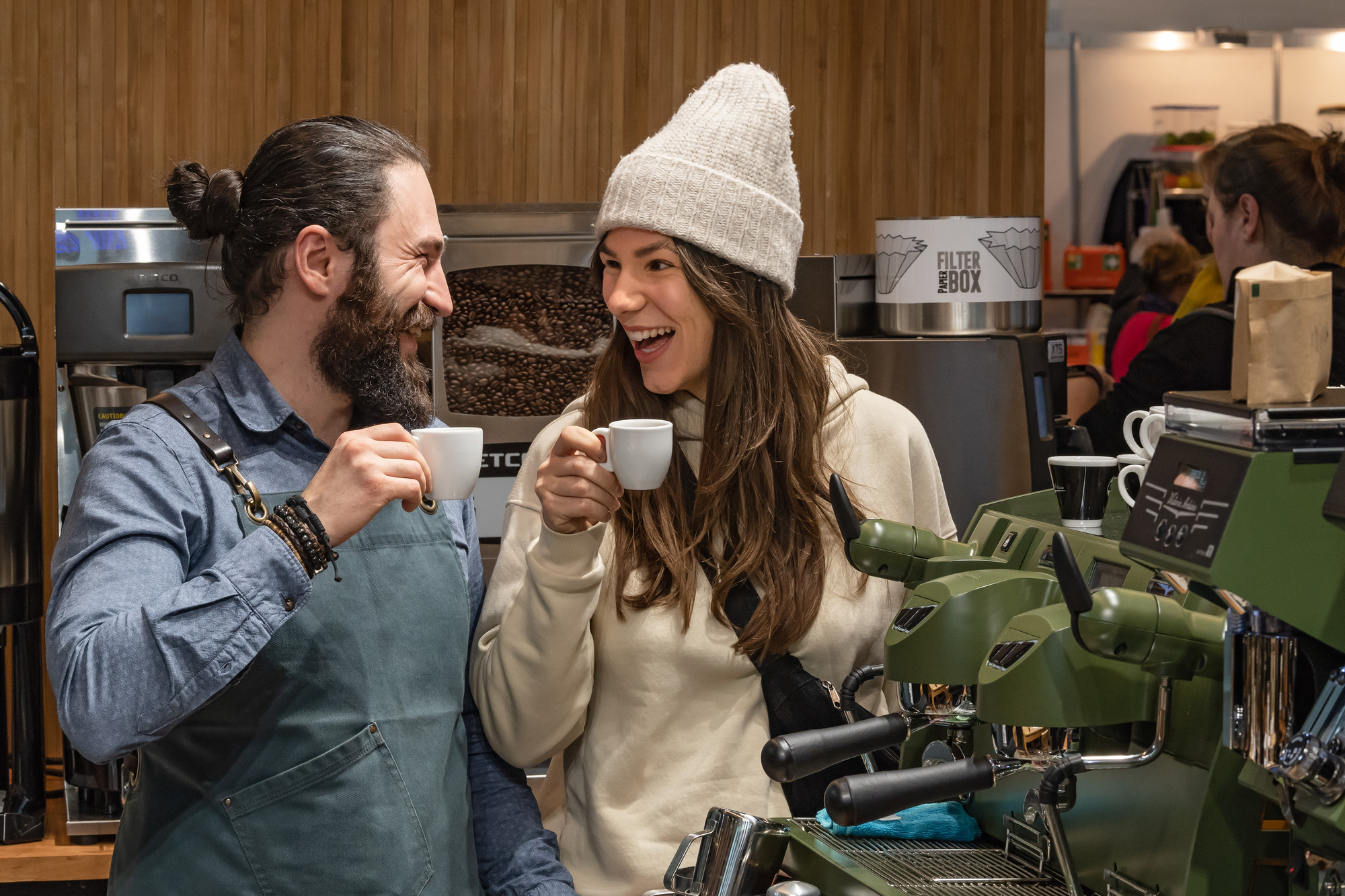 Barista und junge Frau strahlen sich an mit einer Tasse Kaffee in der Hand. Gastro Ivent 2020
