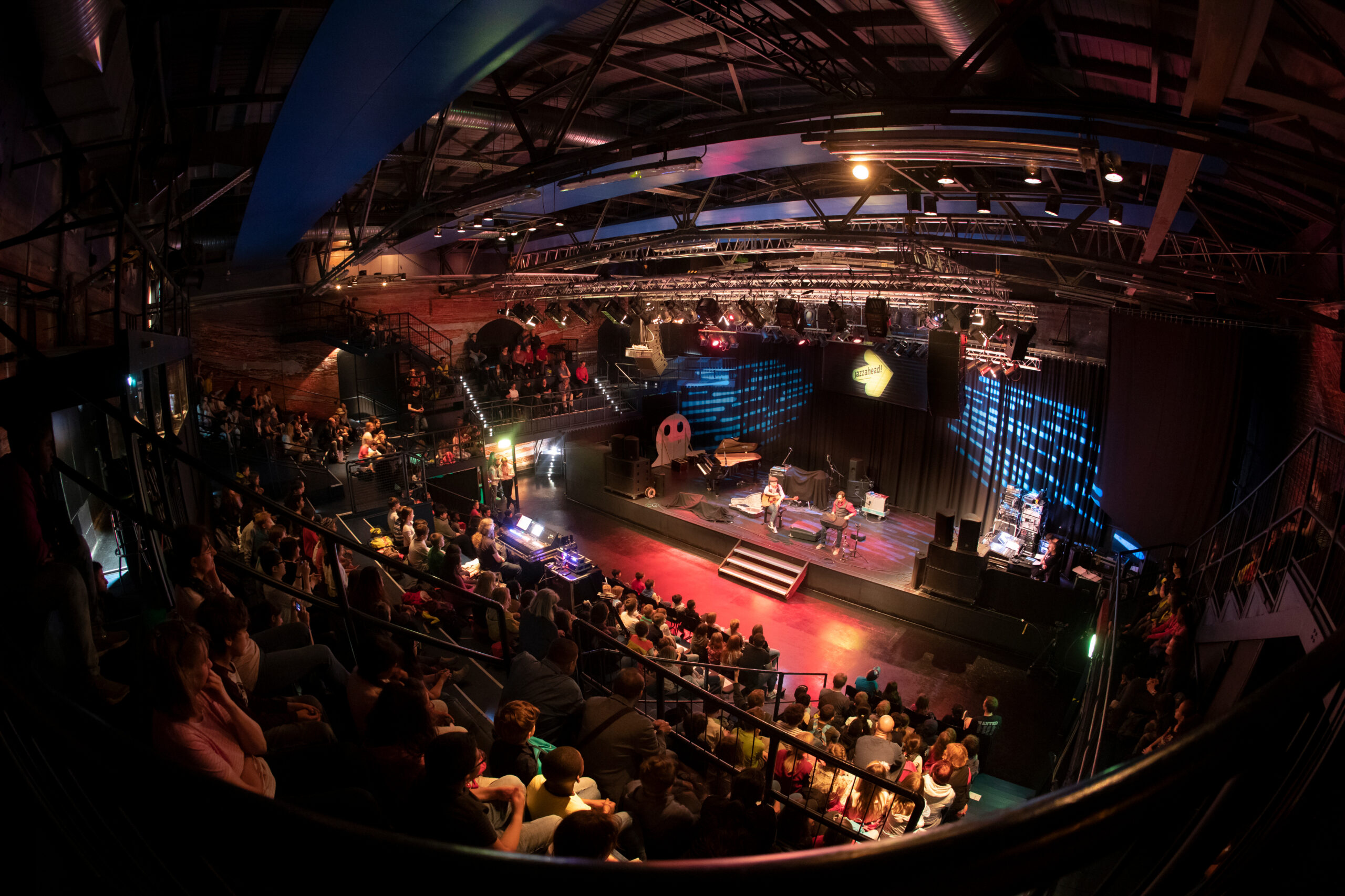 Ein vollbesetztes Theater, in dem die Zuschauer sitzen und eine Live-Aufführung auf der Bühne verfolgen, mit Musikern und farbenfroher Bühnenbeleuchtung, von einer oberen Ebene aus gesehen.